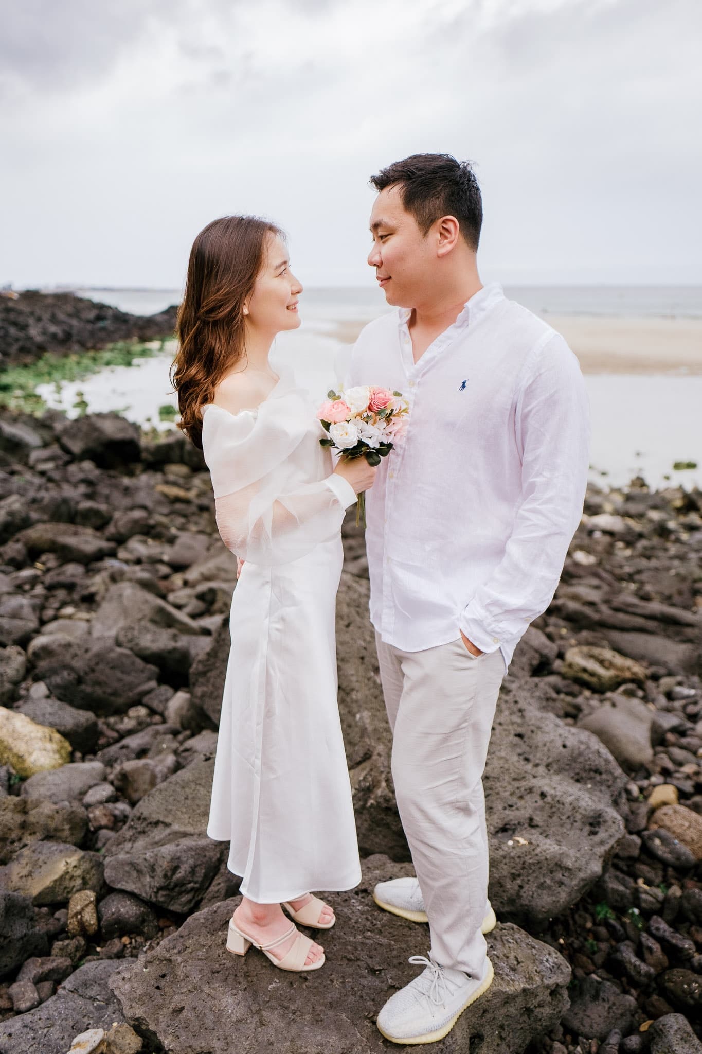 Couple in white on a rocky shoreline, bouquet in hand