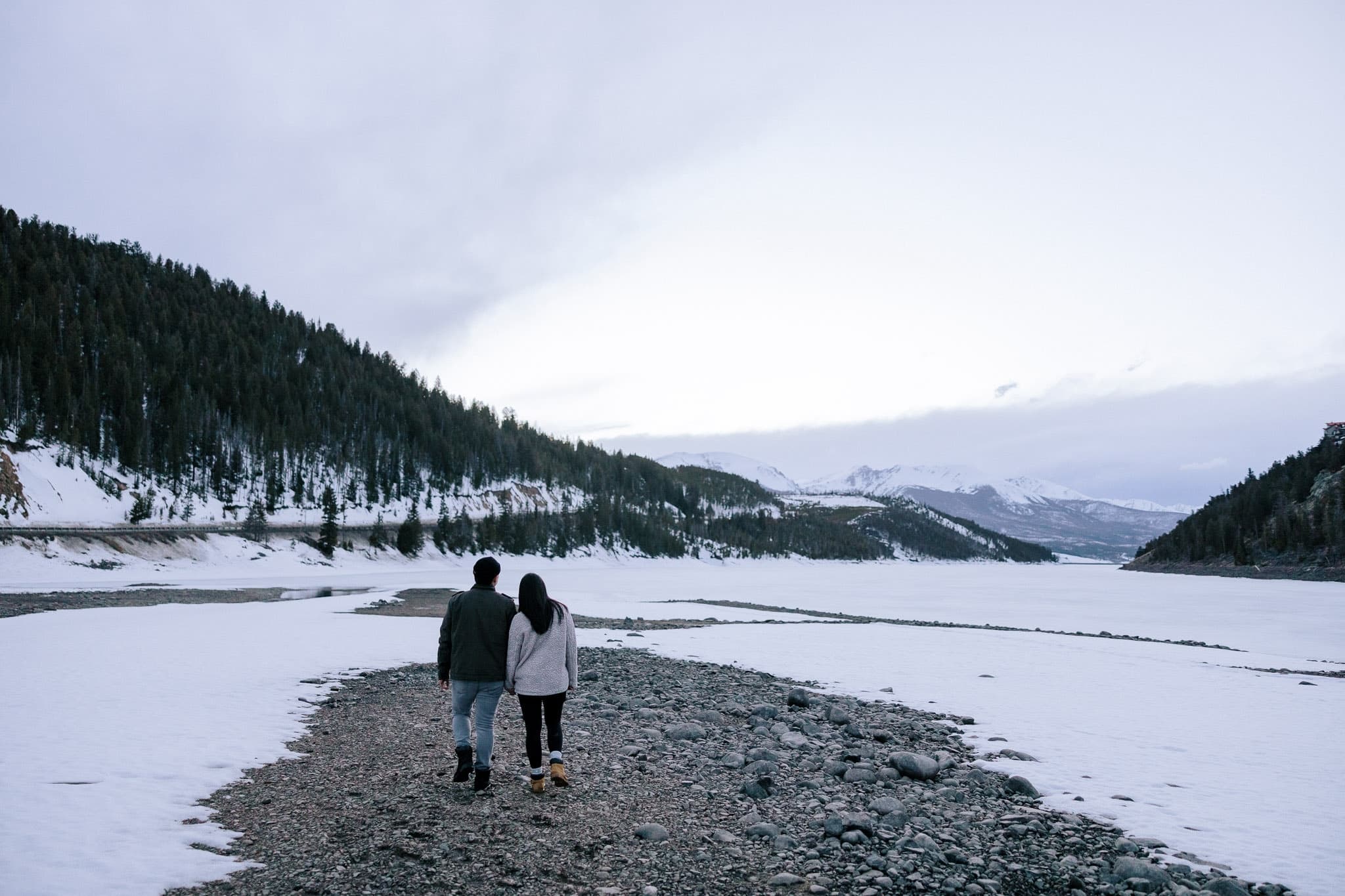 Couple walking along a snowy lakeshore beneath the hills