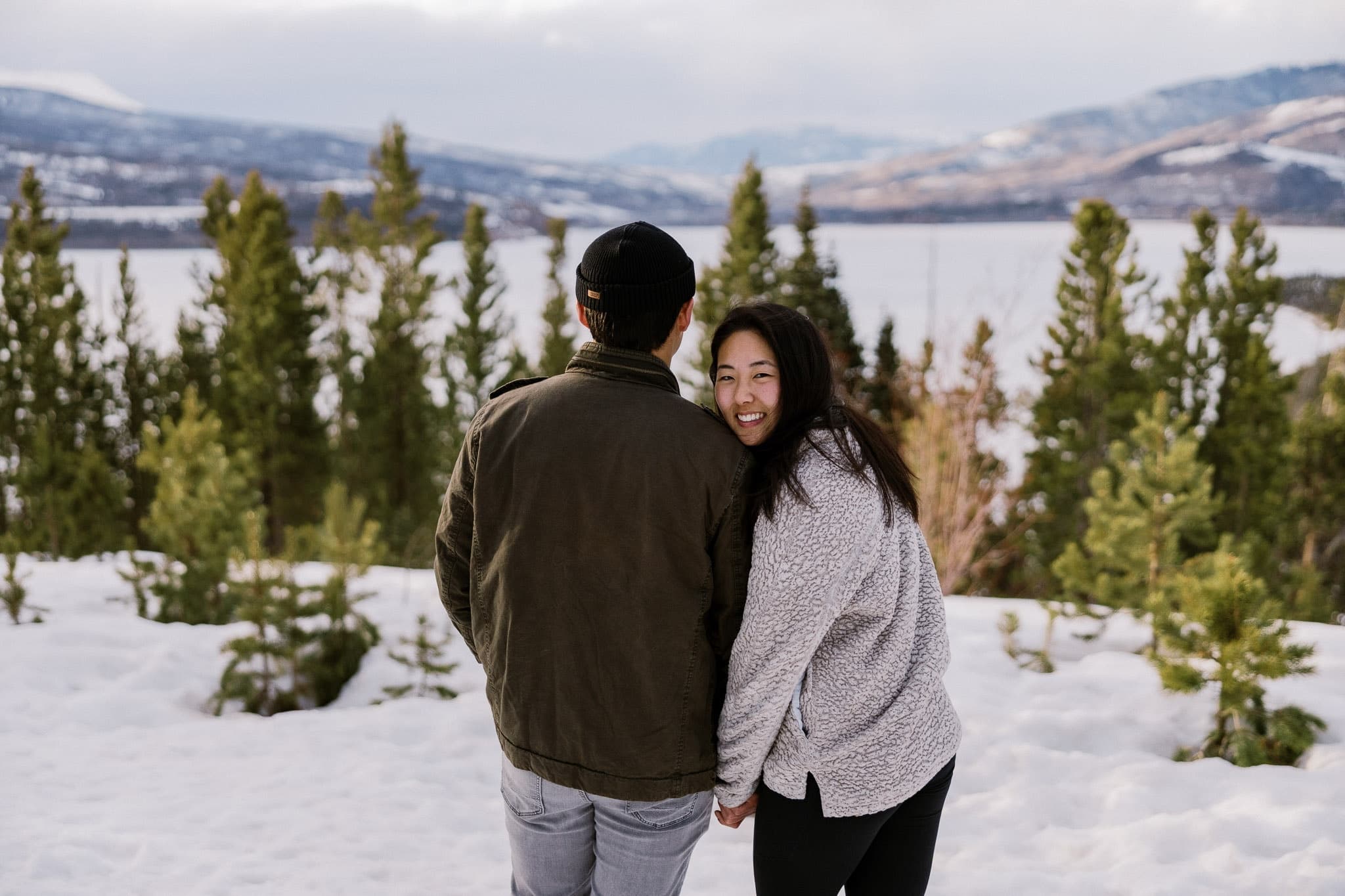 Couple facing the snowy mountains, one glancing back with a smile