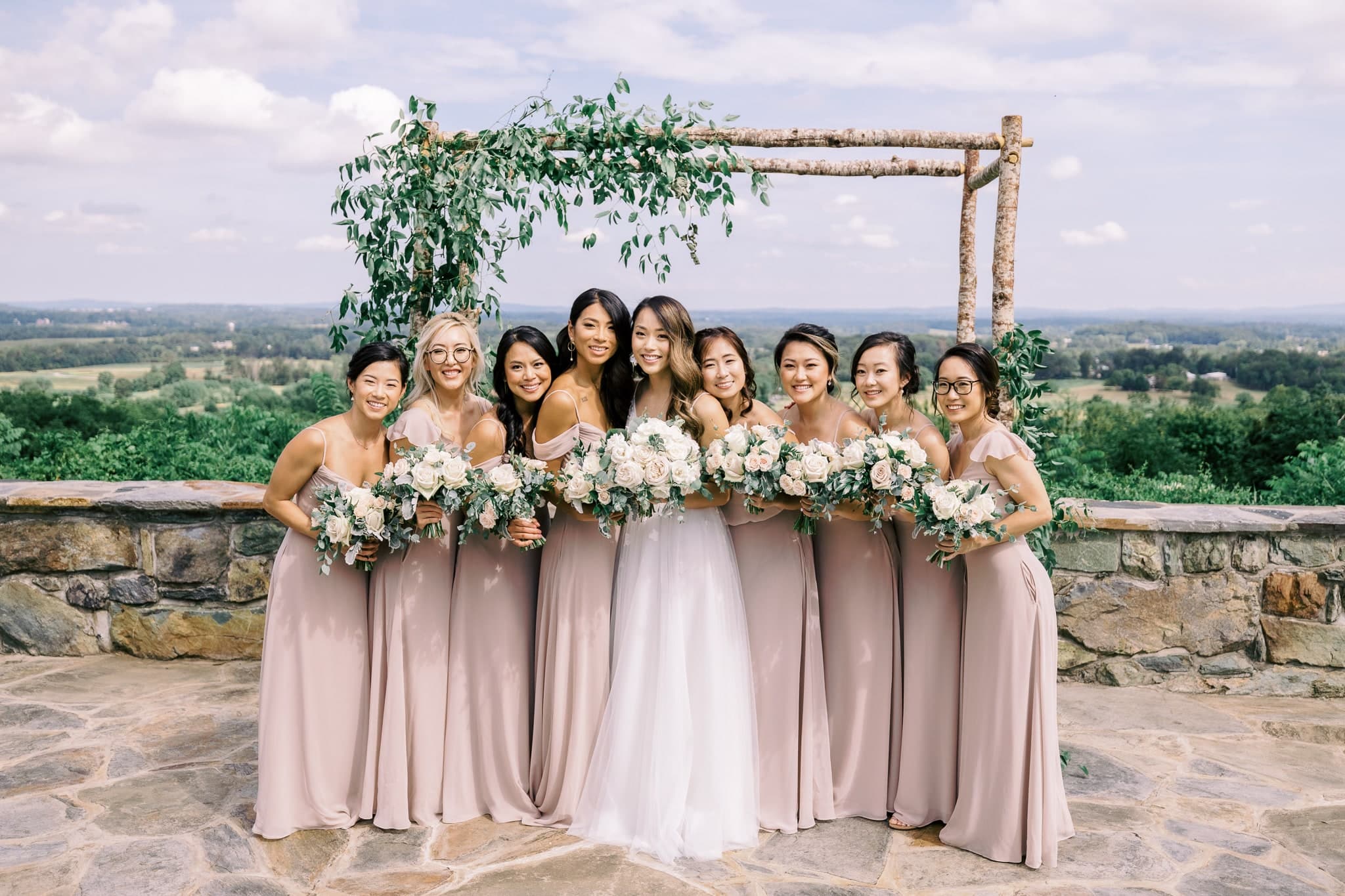 Bride with her bridesmaids at a greenery arbor overlooking the hills